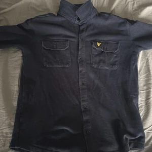 Svart overshirt från Lyle & Scott - Svart overshirt från Lyle & Scott med klassisk krage, två bröstfickor med knappar och den ikoniska gula loggan broderad på ena fickan. Skjortan är i mjuk bomull och har knäppning framtill. Perfekt för en stilren och avslappnad look.