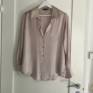 Champagnefärgad silkesblus med knappar - Snygg champagnefärgad skjorta i silkesliknande material med klassisk krage och långa ärmar. Skjortan har en loose fit och guldiga knappar framtill samt vid manschetten. Perfekt för dig som gillar en stilren och lyxig look. Aldrig använd!💗🙌🏼