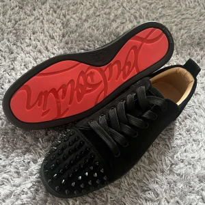 Christian Louboutin svarta sneakers med nitar - Säljer ett par svarta sneakers från Christian Louboutin med ikonisk röd sula och svarta nitar på tån. Skorna har snörning, rund tå och är tillverkade i mocka med läderdetaljer. Kommer med originalkartong, dustbag och äkthetskort.