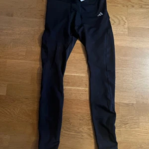 Svarta leggings från Adidas - Svarta leggings från Adidas med klassisk logga på höften. Tight passform och stretchigt material som passar perfekt till träning. Tillverkade i polyester och designade för att sitta skönt hela dagen.