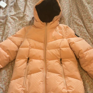 Beige pufferjacka från Nikkie - Säljer en rosa (den är mer rosa i verkligheten) pufferjacka skidjacka från Nikkie i storlek 38. Jackan har huva, två dragkedjeförsedda fickor och en stor logga på bröstet. Insidan är svart och materialet känns mjukt och syntetiskt, perfekt för kalla dagar. Stängs med dragkedja framtill. Använd 2 veckor i sälen. Lite smutsig.