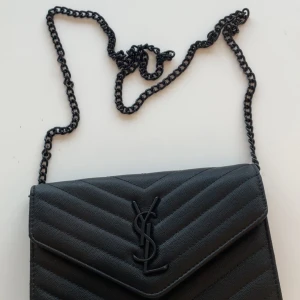 Svart quiltad axelväska från Saint Laurent - Snygg svart axelväska från Saint Laurent med klassiskt YSL-logo framtill. Väskan är quiltad i chevronmönster och har en kedjerem i svart metall. Tillverkad i skinn med flera fack och röd insida. Perfekt för dig som vill ha en stilren och lyxig väska.