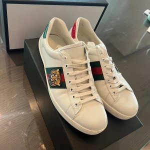 Gucci Ace sneakers  - Gucci Ace sneakers i vitt skinn med klassiska gröna och röda ränder på sidorna, broderad tiger på ena sidan och metallicröd samt metallicgrön detalj på hälen. Snygg och ikonisk modell med snörning och platt sula. Perfekta för dig som vill sticka ut med lyxig streetstyle. Kvitto saknas tyvärr. Kommer i original box 