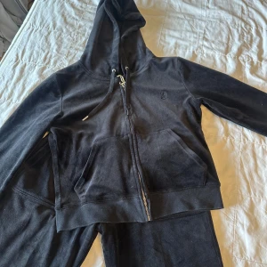 Svart Juicy Couture SET - Svart hoodie från Juicy Couture i mjuk velour med dragkedja, huva och snörning. Klassisk JC-logga broderad på bröstet och fickor framtill. Perfekt för en chill och trendig look💗