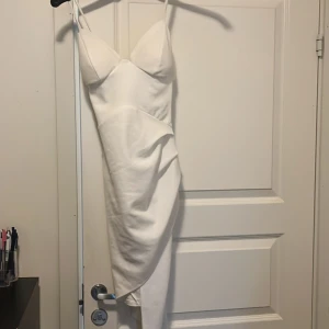Vit asymmetrisk långklänning - Säljer en vit långklänning med smala axelband och figurnära passform. Klänningen har en snygg asymmetrisk nederkant och markerad byst. Perfekt för dig som vill sticka ut med en stilren och modern look.