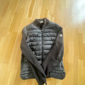 Svart Moncler pufferjacka med stickade ärmar - Snygg svart pufferjacka från Moncler med quiltad framsida och stickade ärmar. Jackan har dragkedja framtill, ståkrage och Moncler-logga på ärmen. Perfekt för dig som vill ha en stilren och sportig look med exklusiv känsla. Kan tyvärr inte garantera äktheten 