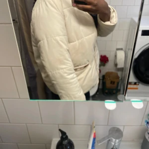 Beige pufferjacka med dragkedja - Säljer en beige pufferjacka med dragkedja framtill och två fickor med snygga detaljer. Jackan har en clean look och är perfekt för dig som gillar en enkel men trendig stil. Långärmad och med normal passform, passar till kyliga dagar.