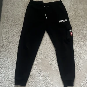 Svarta Hoodrich cargopants med tryck - Svarta mjukisbyxor från Hoodrich med vit logga på benet och fickan. Byxorna har snörning i midjan, elastiska muddar vid benslut och en stor ficka på sidan med tryck. Den har även en ficka med dragkedja intill benet. Mjukisbyxorna är värkligen super mjuka och sköna. Tillverkade i mjuk bomullsblandning för skön passform.