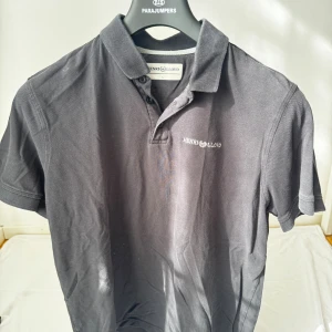 Svart pikétröja från Henri Lloyd - Snygg svart pikétröja från Henri Lloyd i storlek L. Klassisk krage och knappslå framtill, med diskret logga broderad på bröstet. Tillverkad i mjuk bomull som känns skön mot huden. Perfekt för en chill och stilren look.