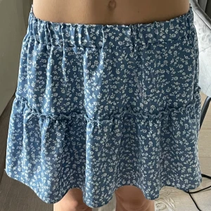 Blå blommig volangkjol - Supersöt blå kortkjol med små vita blommor och volangdetalj. Perfekt för dig som gillar en feminin och somrig stil. Resår i midjan gör den bekväm och enkel att bära.