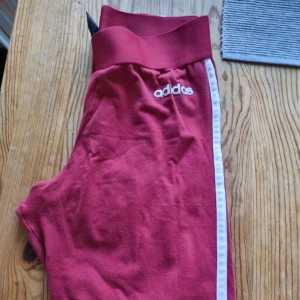 Röda Adidas mjukisbyxor med vit rand - Röda mjukisbyxor från Adidas med vit sidrand och broderad logga på höften. Byxorna har bred resår i midjan och är tillverkade i mjukt material som passar perfekt för chill eller träning.