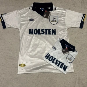 Tottenham Hotspur vit fotbollströja Umbro - Snygg Tottenham Hotspur fotbollströja från Umbro i vit med subtila diagonala ränder och mörkblå detaljer. Klassisk krage med klubbmärke och Holsten-tryck på bröstet. Kortärmad och tillverkad i glansigt polyester-material, perfekt för match eller samling.