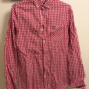 Rutig röd skjorta från Lyle & Scott - Röd och vit rutig skjorta från Lyle & Scott med klassisk krage, bröstficka och knappar framtill. Skjortan har lång ärm och ikonisk gul logga på fickan. Perfekt för dig som gillar en stilren men ändå färgstark look.