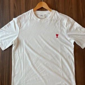 Ami Paris T-Shirt - Vit t-shirt från Ami Alexandre Mattiussi med klassisk passform och rund hals. På bröstet finns en broderad röd logga med hjärta och bokstaven A. Tillverkad i mjuk bomull, perfekt för en clean och stilren look. Den är lite oversized