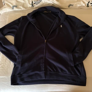 Mörkblå ziptröja från Polo Ralph Lauren - Mörkblå långärmad ziptröja från Polo Ralph Lauren med klassisk vit broderad logga på bröstet. Tröjan har hel dragkedja framtill, två fickor och är tillverkad i mjuk bomull. Perfekt för lager-på-lager och chill vardagsstil.