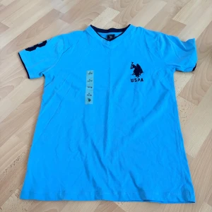 Blå t-shirt från U.S. Polo Assn. - Snygg blå t-shirt från U.S. Polo Assn. med mörkblå detaljer vid krage och ärmslut. T-shirten har v-ringning och tryckt logga på bröstet. Tillverkad i 100% bomull, perfekt för en avslappnad och sportig look.
