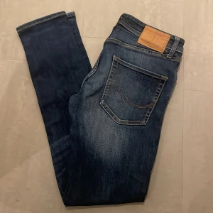 Jack & Jones Slim Fit Tim Jeans - Mörkblå jeans från Jack & Jones i modellen Slim Fit Tim. Jeansen har snygga slitningar och kontrastsömmar, klassisk femficksdesign och läderpatch bak i midjan. Tillverkade i stretchig denim för en bekväm passform.