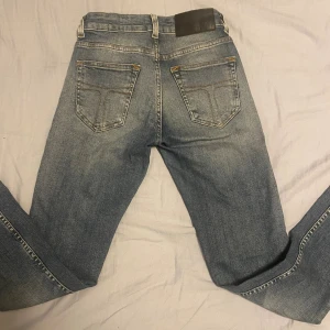Blå jeans från Tiger of Sweden små slitningar - Snygga blå jeans från Tiger of Sweden med klassisk femficksdesign och slitna detaljer framtill. Jeansen har raka ben och normal passform, med snygga kontrastsömmar och läderpatch bak i midjan. Perfekta för en avslappnad och trendig look.