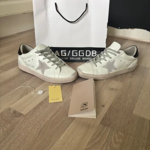 Golden Goose Superstar sneakers vit/grå - Golden Goose Superstar sneakers i vitt skinn med grå mockadetaljer och stjärna på sidan. Grå snörning, svart hälparti i skinn och klassisk platt sula. Snygg och ikonisk modell med cool vintagekänsla, perfekt för dig som gillar streetstyle.