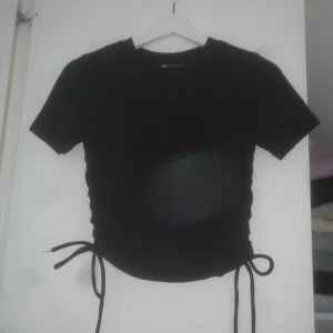 Svart snörad topp  - Svart kortärmad topp från Zara med snörning i sidorna som ger en snygg, justerbar passform. T-shirten har rund halsringning och är tillverkad i mjukt bomullsmaterial. Perfekt att styla med jeans eller kjol för en trendig look.