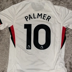 Chelsea Palmer 10 bortatröja Nike S - Chelsea FC bortatröja med Palmer 10-tryck. Vit med svarta och röda detaljer, tunna vertikala ränder framtill och broderat klubbmärke. Tillverkad i lätt och ventilerande polyester med Dri-FIT-teknologi. Klassisk passform och korta ärmar.