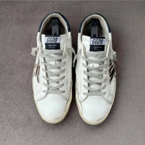 GGDB vita sneakers med zebramönster - Snygga vita sneakers från GGDB med zebramönstrad stjärna på sidan och mörkblå häl. Skorna har snörning, platt sula och är tillverkade i skinn. Perfekta för dig som gillar en unik och trendig look.