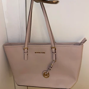 Rosa Michael Kors axelväska  - Snygg rosa axelväska från Michael Kors i skinn med guldiga detaljer och MK-hänge. Väskan har två långa axelband, dragkedja upptill och flera fack inuti för smart förvaring. Perfekt storlek för att få plats med allt du behöver.