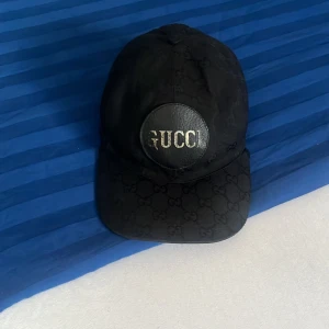 Svart keps från Gucci med logga - Svart keps från Gucci med klassiskt GG-monogram över hela kepsen och en rund svart patch framtill med silvrig GUCCI-logga. Justerbar rem baktill för perfekt passform. Snygg och stilren accessoar som lyfter vilken outfit som helst.