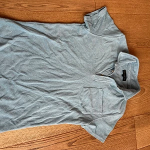 Ljusblå frotté t-shirt från LAGENCY - Säljer en ljusblå t-shirt från LAGENCY i mjuk frotté. T-shirten har korta ärmar, bröstficka och knappar vid kragen för en chill look. Perfekt för dig som gillar en avslappnad stil med lite extra känsla.