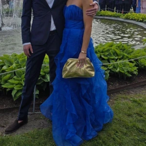 Blå långklänning med volanger - Säljer en fantastisk blå långklänning med volanger och axelbandslös design. Klänningen har en vacker omlottkänsla upptill och flera lager av transparent tyg som ger ett riktigt drömmigt intryck. Perfekt för bal!! Sitter snyggt och ger en elegant siluett. Fick mycket komplimanger. 