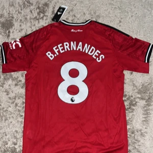 Manchester United Fernandes tröja L - Säljer en officiell Manchester United fotbollströja från Adidas med B. Fernandes och nummer 8 på ryggen. Tröjan är röd med svarta och vita detaljer, broderat klubbmärke och sponsorlogga på bröstet. Tillverkad i lätt och ventilerande polyester.
