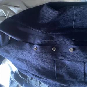 Mörkblå stickad cardigan med knappar - Snygg mörkblå stickad cardiganmed klassisk krage och stora knappar framtill. Jackan har en rak passform och praktiska fickor. Perfekt för dig som gillar stilrena och tidlösa plagg.