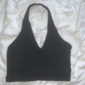 Svart halterneck topp  - Svart halterneck topp från ASOS med djup v-ringning och croppad passform. Toppen är ärmlös och har en stilren, minimalistisk look som passar perfekt till jeans eller kjol. Tillverkad i mjukt material som känns skönt mot huden.