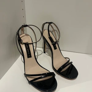 Svarta klackade sandaler från Zara - Säljer ett par stilrena svarta sandaler från Zara med smala remmar och hög klack. Skorna har öppen tå och justerbart spänne runt vristen. Perfekta för dig som gillar en elegant och minimalistisk look. Materialet är glansigt syntet.