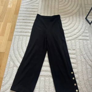 Svarta wide byxor med knappar - Snygga svarta byxor med wide fit och hög midja. Byxorna har dekorativa silvriga knappar längs ena benet och är tillverkade i ett mjukt bomullsmaterial. Perfekt för dig som gillar stilrena och trendiga detaljer.