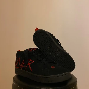 DC Shoes x Slayer svarta sneakers - Svarta sneakers från DC Shoes i samarbete med Slayer. Skorna har röd Slayer-logga broderad på sidan och tungan samt pentagramdetalj framtill. Grova svarta skosnören och röd hälflik. Ovandel i mocka och syntet, platt sula och rund tå.