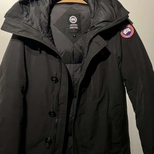 Svart dunjacka från Canada Goose - Svart dunjacka från Canada Goose med klassisk patch på ärmen. Jackan har huva, dragkedja och stora knappar framtill. Fodrad för extra värme och har flera fickor. Perfekt för kalla vinterdagar och har en stilren look som funkar till allt. Kan även tänka mig gå ner i pris om det är snabb köp☺️