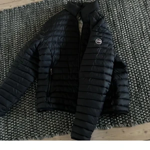 Colmar jacka -  Storlek 50 å passar dig som är 180 typ. Snygg svart pufferjacka från Colmar med horisontella quiltade paneler och hög krage. Jackan har dragkedja framtill och två sidofickor med dragkedja. Perfekt för kyliga dagar och har en stilren look med diskret logga på bröstet.