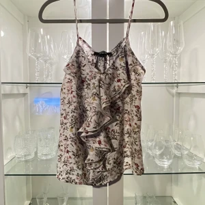 Blommig linnetopp med volang Bik Bok XS - Supersöt linnetopp från Bik Bok i ljusrosa med blommigt mönster i rött, gult och grönt. Tunt, glansigt material med smala axelband och en stor volang framtill som ger en extra touch. Perfekt för dig som gillar romantisk och feminin stil.