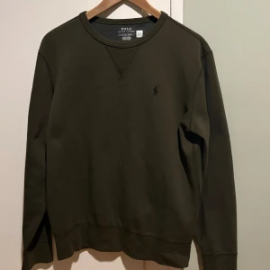 Olivgrön sweatshirt från Polo Ralph Lauren - Olivgrön sweatshirt från Polo Ralph Lauren med klassisk rund hals och broderad logga på bröstet. Tröjan har långa ärmar och ribbade muddar vid ärmslut och nederkant. Tillverkad i mjuk bomull för skön komfort och stilren look.