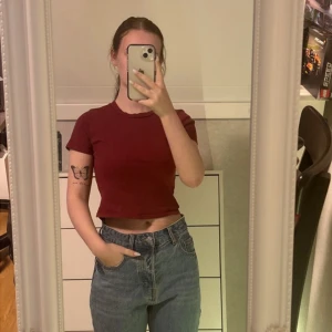 Mörkröd croppad ribbad topp - Säljer en mörkröd croppad topp med korta ärmar och rund hals. Toppen är ribbad och har en enkel, stilren design som passar perfekt till jeans eller kjol. Materialet känns mjukt och stretchigt, vilket gör den bekväm att bära hela dagen.