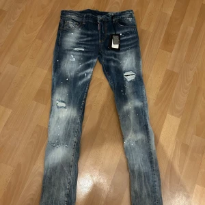 Dsquared2 blå slim jeans med slitningar - Snygga blå jeans från Dsquared2 i slim fit med coola slitningar, färgstänk och distressed detaljer. Klassisk femficksmodell med Dsquared2-logga på baksidan. Jeansen har en modern tvätt och är tillverkade i bomull. Oanvända.