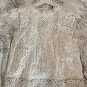 Vit spetsblus från ONLY - Superfin vit blus från ONLY med detaljerad spets och broderade kanter. Blusen är ärmlös med hög hals och har vackra volangdetaljer längs axlarna. Perfekt för dig som gillar romantisk och feminin stil.