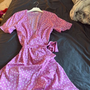 Rosa blommig omlottklänning - Superfin rosa omlottklänning från ONLY med vita små blommor. Klänningen har korta ärmar, knyt i midjan och volangdetaljer längs nederkanten. Perfekt för varma dagar och ger en söt och somrig vibe.