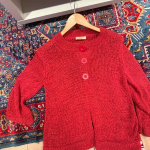 Röd stickad kofta från Oui - Säljer en röd stickad kofta från Oui med tre stora matchande knappar framtill. Koftan har en rund halsringning och är långärmad, perfekt för lager på lager. Den har en loose passform och är riktigt skön att slänga på sig över en t-shirt. Tröjan passar allt från xs-L beroende på hur man vill att den ska sitta