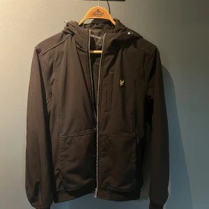 Svart vindjacka från Lyle & Scott - Svart vindjacka från Lyle & Scott med huva och dragkedja framtill. Jackan har två stora fickor, ribbade muddar och en gul broderad logga på bröstet. Perfekt för blåsiga dagar och har en clean, sportig look som funkar till jeans eller joggers.