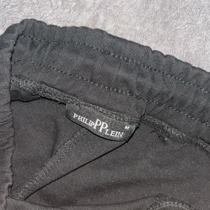 Svarta mjukisbyxor från Philipp Plein - Svarta mjukisbyxor från Philipp Plein med elastisk midja och snörning. Byxorna har dragkedjor med märkets logga och detaljer i blankt material vid fickorna. Snygg och modern look med coola detaljer och bekväm passform.