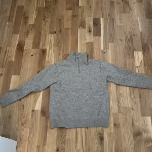 Grå merinoulltröja från Vaillent - Mysig grå stickad tröja från Vaillent i mjuk merinoull. Tröjan har hög krage med half zip-dragkedja och långa ärmar. Perfekt för lager-på-lager och chill dagar. Enkel och stilren design som funkar till det mesta.