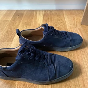 Mörkblå mockasneakers från Louboutin - Snygga mörkblå sneakers i mocka från Christian Louboutin med klassisk röd sula och rund tå. Skorna har snörning och en stilren design som passar till det mesta. Insidan är ljusbeige och sulan har Louboutins ikoniska signatur.