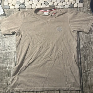 Beige Moncler t-shirt i bomull - Snygg beige t-shirt från Moncler med diskret logga på bröstet. Klassisk rund hals med röd, vit och svart randig insida. Tillverkad i mjuk bomull och har korta ärmar. Perfekt för dig som gillar stilrena och exklusiva plagg.